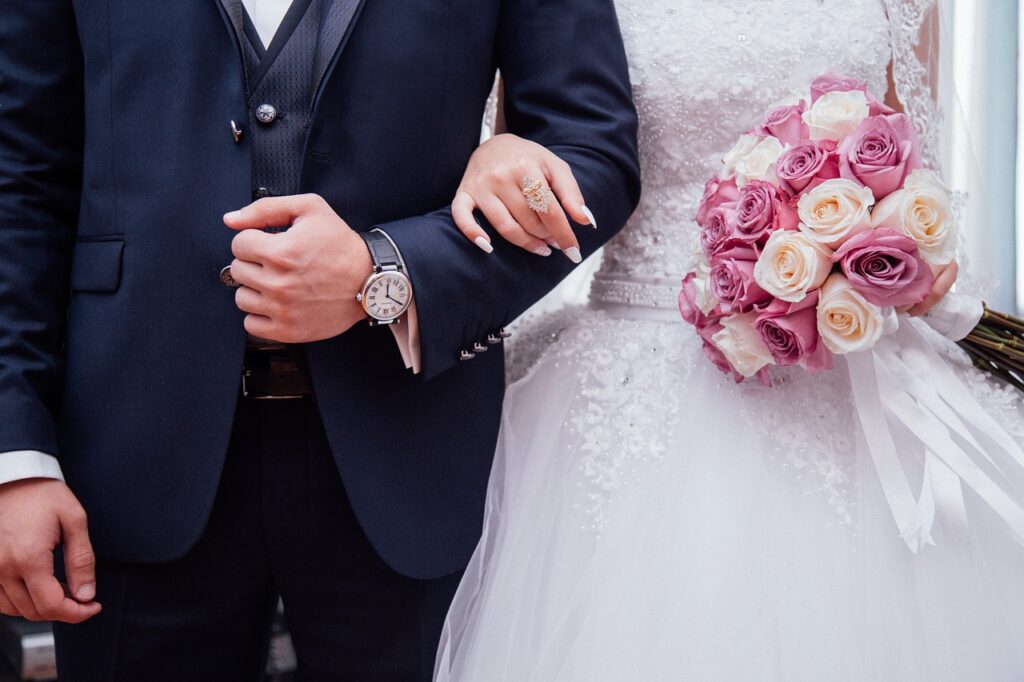Deux mariés se tiennent. L'homme a un costume avec une montre et sa future femme tient un bouquet de fleurs.