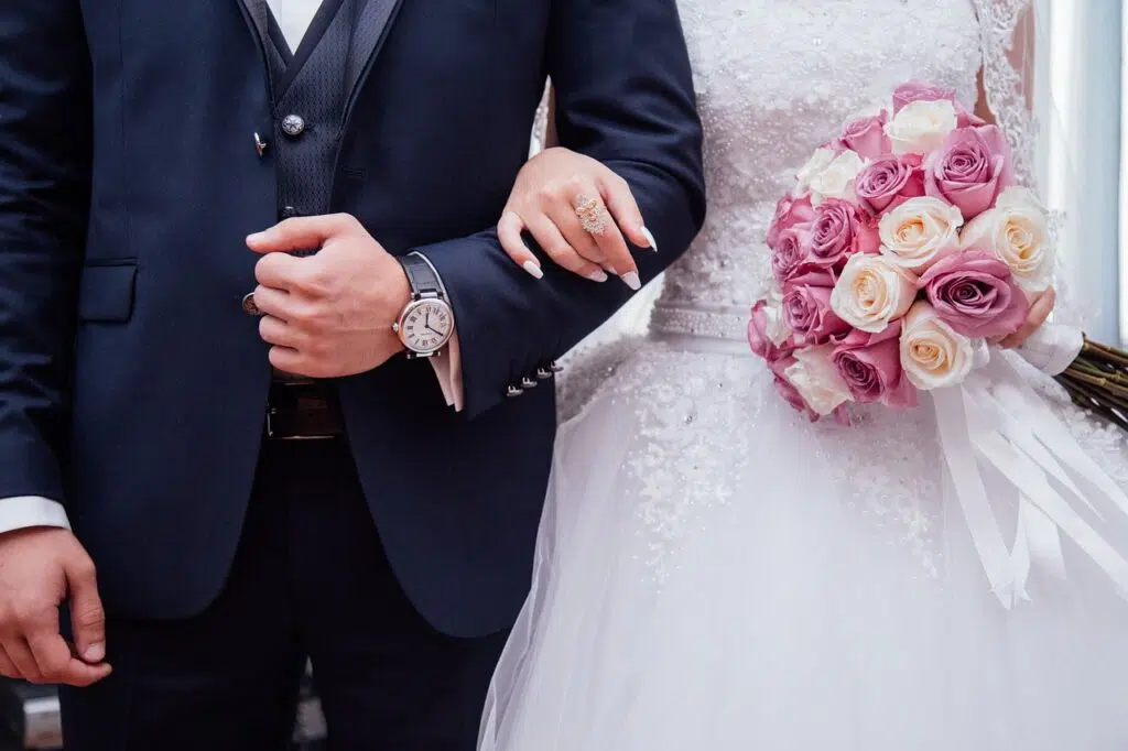 Deux mariés se tiennent. L'homme a un costume avec une montre et sa future femme tient un bouquet de fleurs.