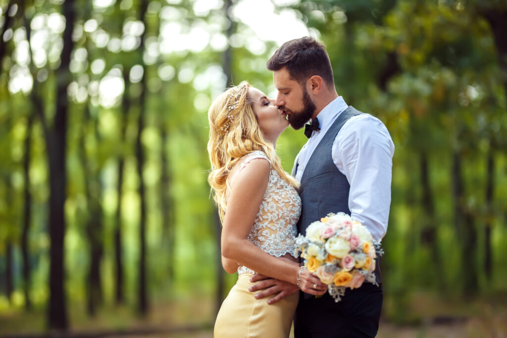 Un couple de mariés s'embrassant tendrement dans un cadre naturel, entouré d'arbres verts. La mariée porte une robe blanche avec des détails en dentelle et tient un bouquet de fleurs colorées, tandis que le marié porte une chemise blanche avec un nœud papillon noir et un gilet gris.