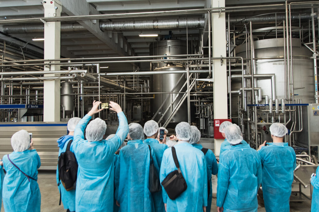 La photo montre un groupe de personnes portant des blouses bleues, des bonnets et des masques, probablement lors d'une visite industrielle. Ils prennent des photos et observent une installation complexe de machines et de tuyauterie en métal, typique d'une usine de transformation ou d'un laboratoire de production.