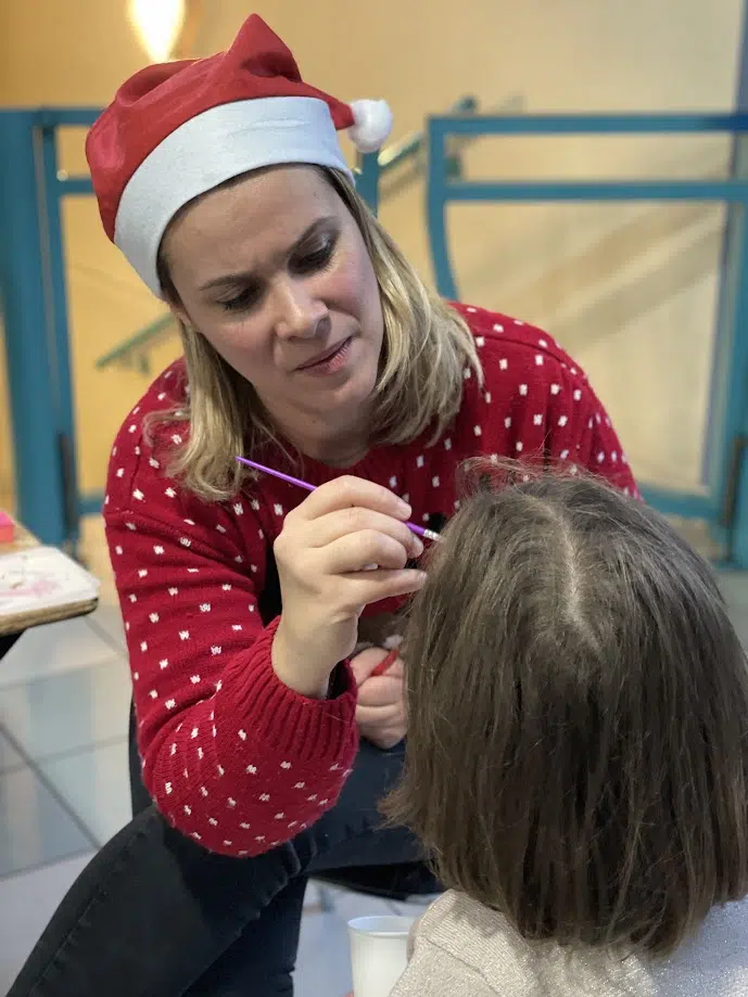 Une femme portant un bonnet de Noël et un pull rouge à motifs blancs est en train de maquiller le visage d'un enfant. Elle est concentrée sur son travail, tenant un pinceau dans la main. L'enfant, dont on voit l'arrière de la tête, est assis calmement pendant que la femme applique le maquillage. La scène se déroule à l'intérieur, avec un fond montrant des rampes d'escalier bleues.