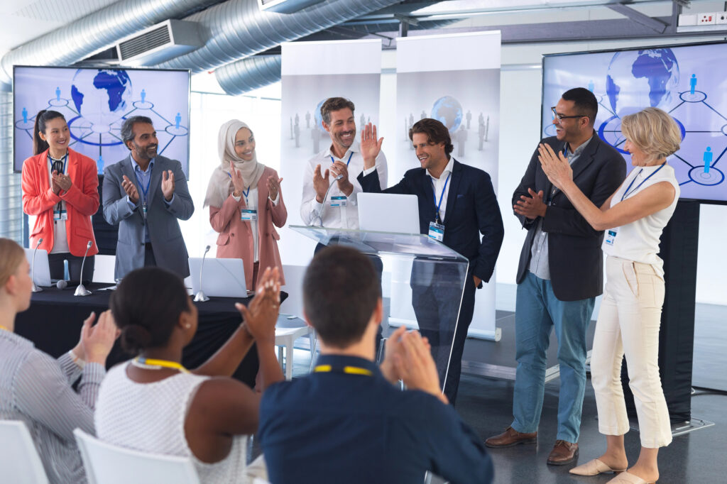 Cette photo montre un groupe diversifié de personnes sur une scène, applaudissant un homme debout derrière un pupitre avec un ordinateur portable. L'événement est une conférence ou une présentation professionnelle, avec des écrans en arrière-plan affichant des graphiques et des connexions globales.