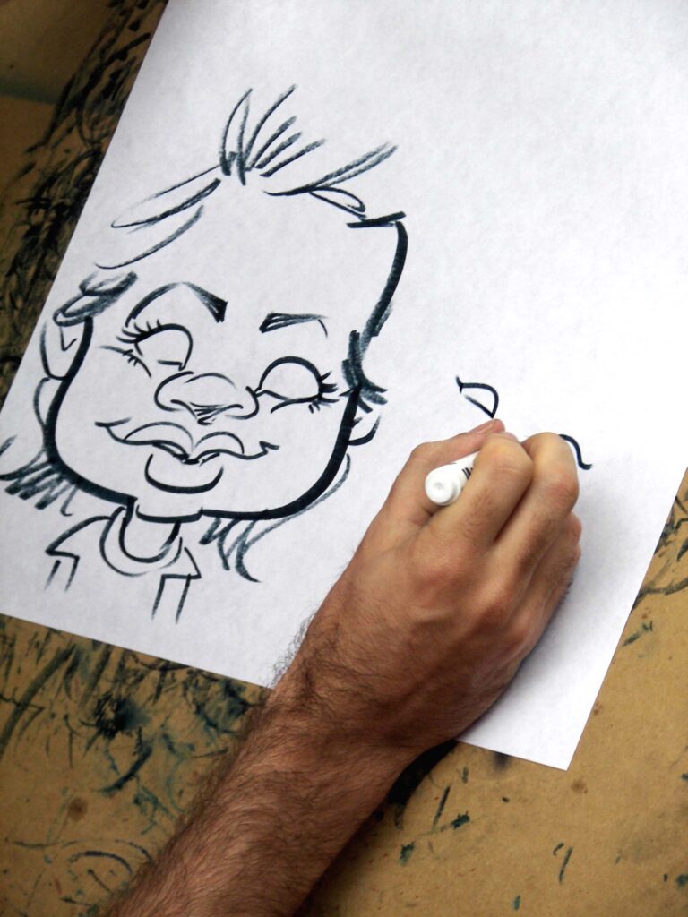 Une main d'artiste est en train de dessiner une caricature amusante d'une personne avec un marqueur noir sur une feuille de papier blanc. Le dessin montre un visage exagéré avec des traits faciaux distinctifs, y compris un grand sourire et des yeux fermés. Le papier est posé sur une surface de travail tachée d'encre. Caricature.