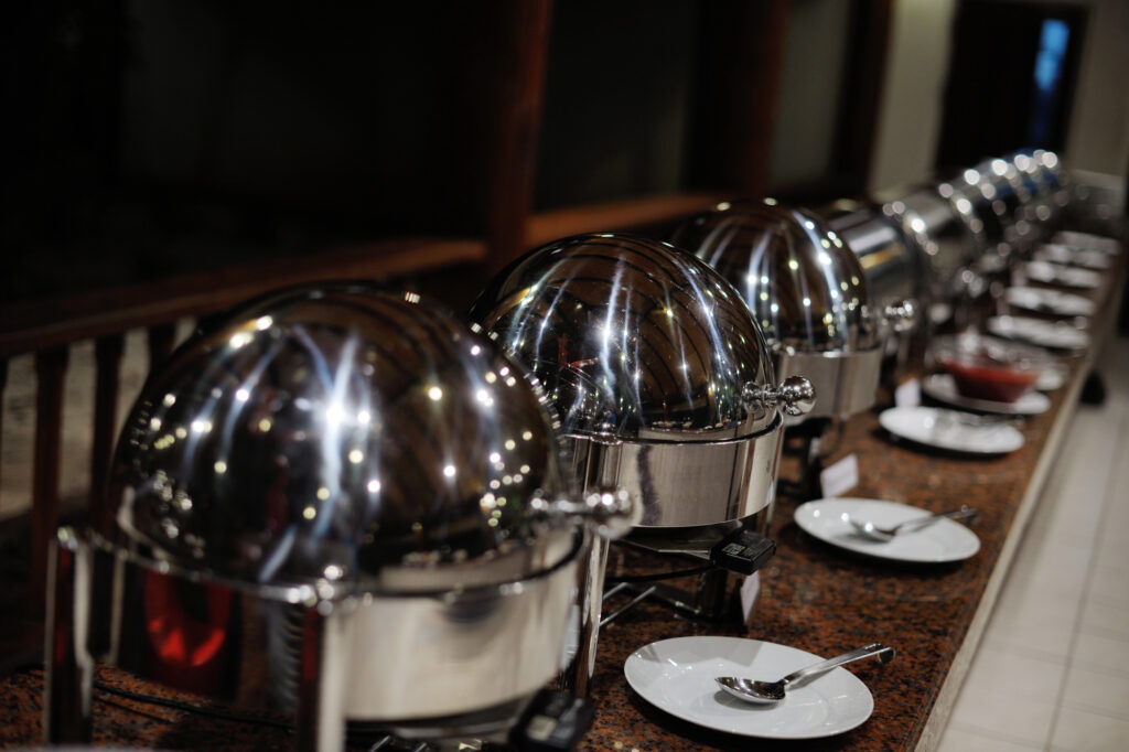 Une rangée de chafing dishes en acier inoxydable, alignés sur un buffet en bois poli, sont prêts à être utilisés pour un service de restauration. Devant chaque plat, des assiettes blanches et des cuillères sont disposées de manière ordonnée, prêtes pour les convives. Les couvercles des plats reflètent les lumières environnantes, créant une ambiance élégante et formelle.