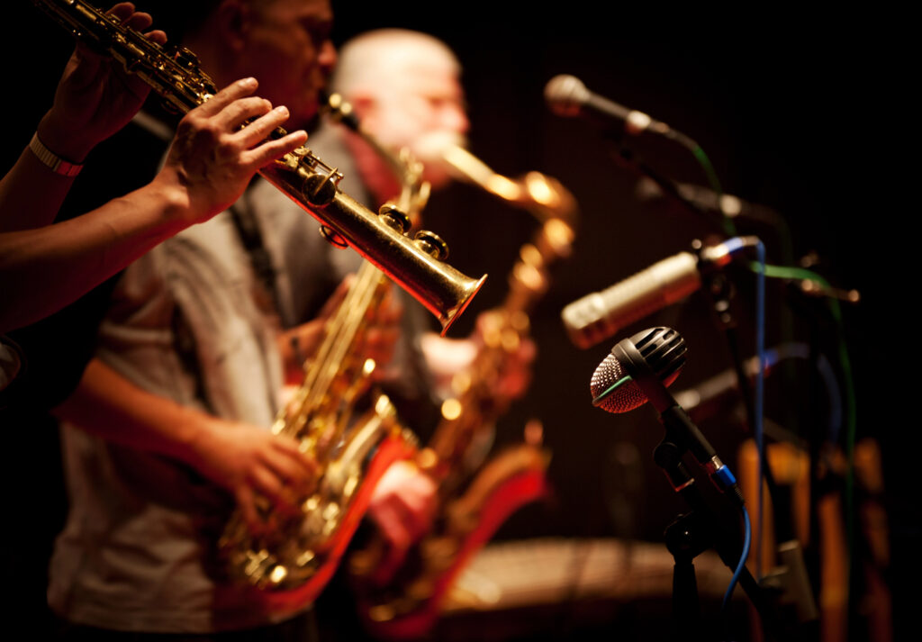 Un groupe de musiciens joue du saxophone sur scène, capturé en gros plan. Les instruments dorés brillent sous les lumières, tandis que des microphones sont positionnés à l'avant, prêts à capter la performance. L'arrière-plan est sombre, mettant en valeur l'intensité et la concentration des musiciens pendant leur prestation.