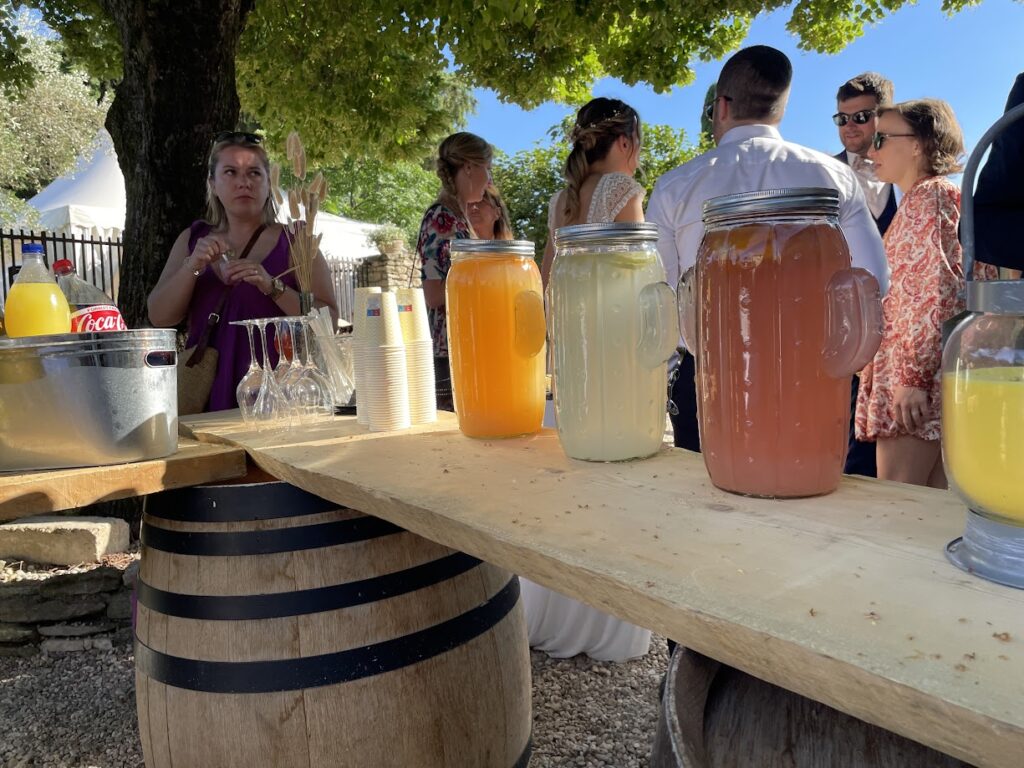 Un bar en plein air est installé sur des tonneaux en bois, servant des boissons fraîches dans de grandes carafes en verre remplies de jus colorés. Des gobelets en plastique sont empilés à côté, prêts à être utilisés par les invités qui se tiennent autour, profitant de l'événement en plein air. L'ombre des arbres au-dessus et le ciel bleu clair en arrière-plan créent une ambiance estivale et décontractée.