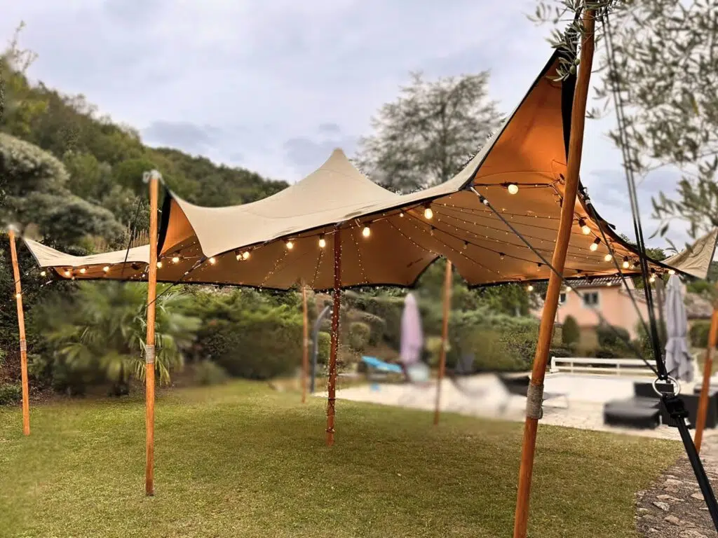 Photo d'une tente nomade montée en extérieur sur une pelouse, éclairée par des guirlandes lumineuses suspendues sous la toile beige. La tente est soutenue par des poteaux en bois et offre un style élégant et naturel, idéal pour un événement en plein air. L'arrière-plan montre un jardin verdoyant et des collines, donnant une ambiance paisible et accueillante pour une réception ou une fête.