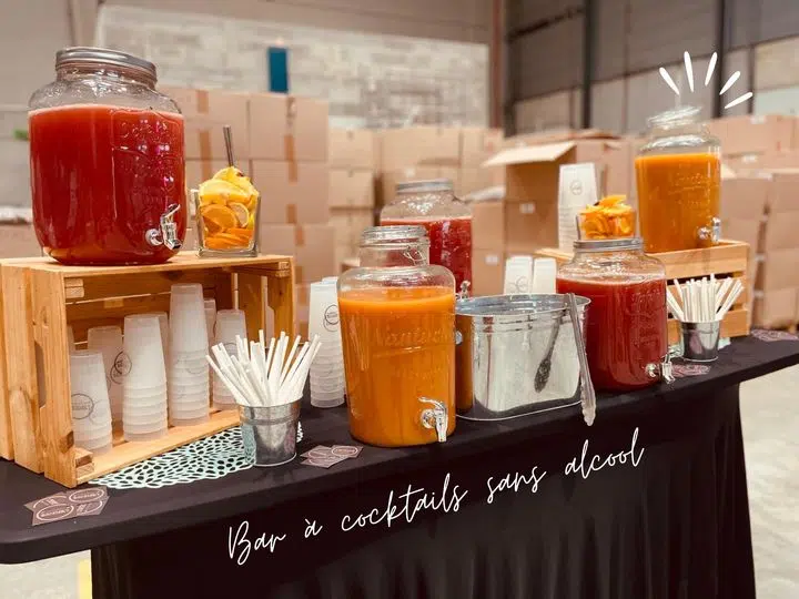 Photo d'un bar à cocktails sans alcool. Sur une table noire, plusieurs grands bocaux en verre remplis de cocktails colorés (rouge, orange, jaune) sont posés, certains sur des caisses en bois. À côté des bocaux, des gobelets en plastique empilés, des tranches d'orange séchées pour la décoration et des pailles blanches sont disposés pour le service. Un seau en métal contenant une louche se trouve au centre, et l'ambiance est industrielle avec des cartons en arrière-plan.